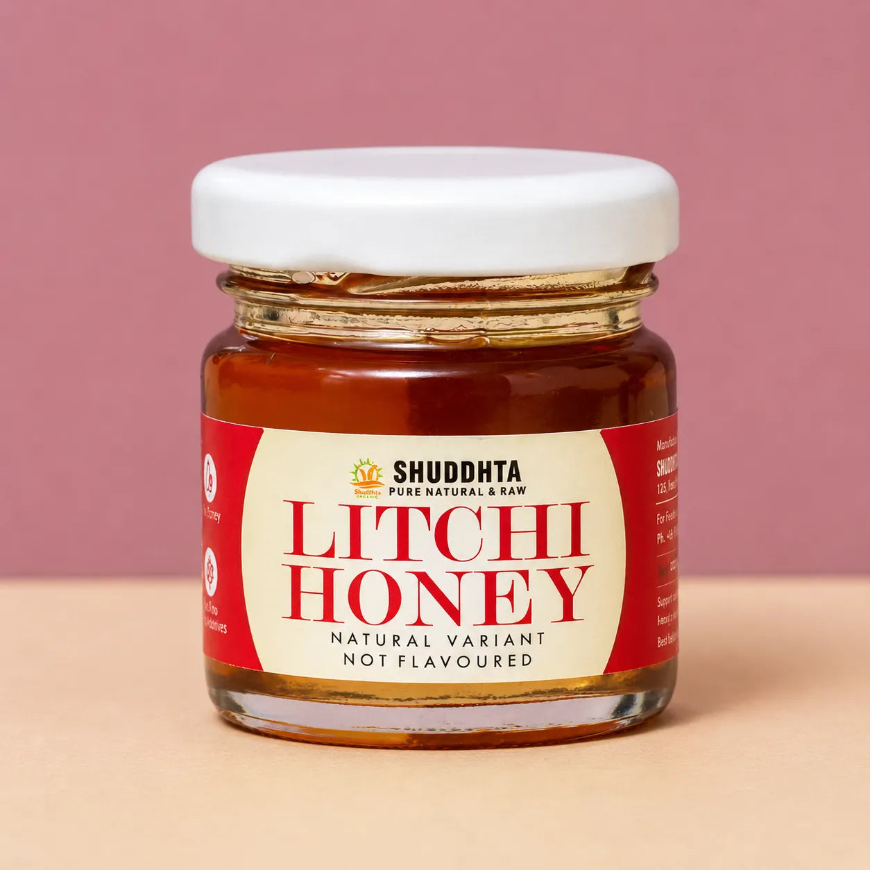 A glass jar of Shuddhta Litchi Honey with a white lid and a cream and red label, set against a solid dusty pink background and a light beige surface.