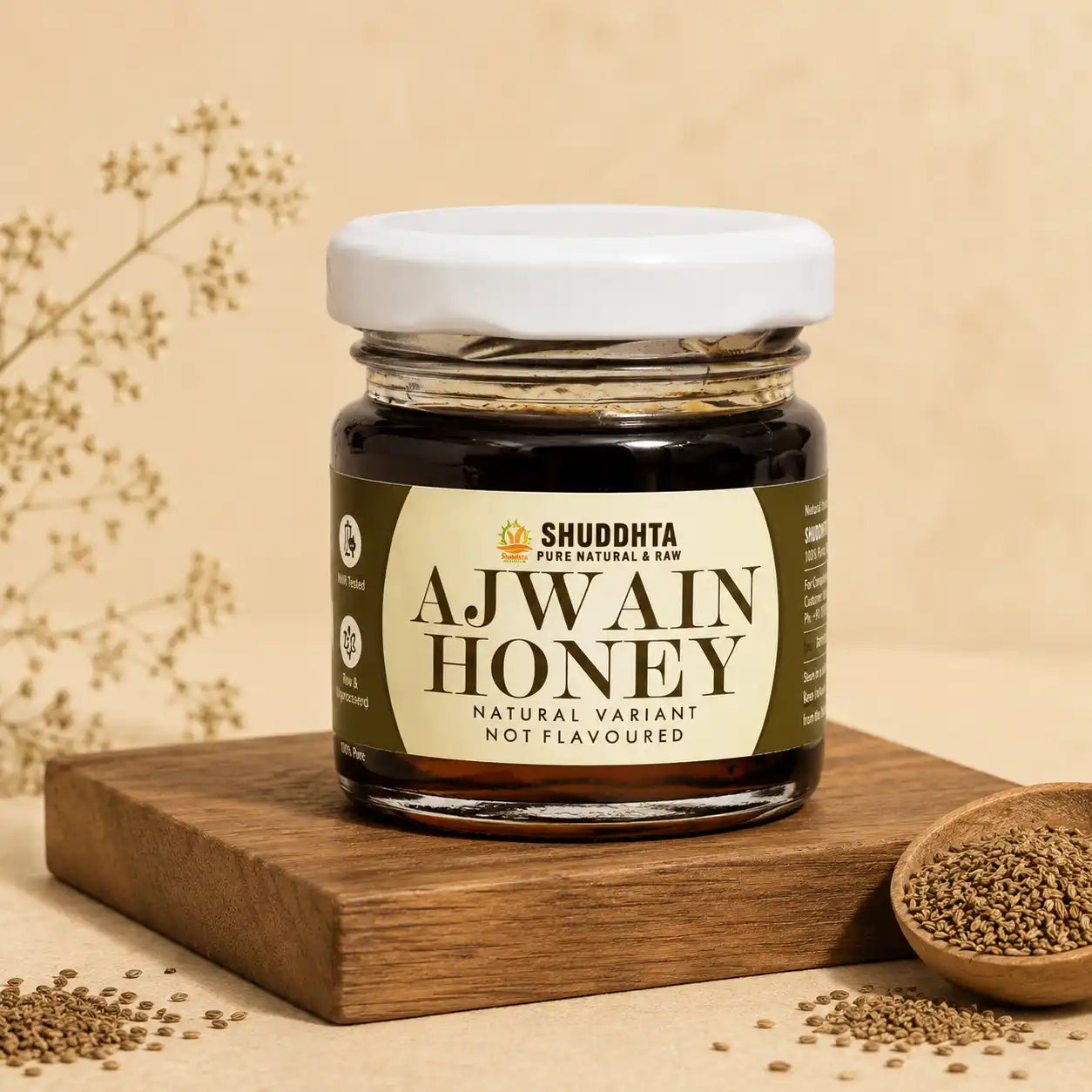 A jar of Shuddhta Ajwain Honey displayed on a wooden block with scattered seeds and a small wooden spoon of seeds on a beige background.