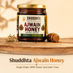 A jar of Shuddhta Ajwain Honey displayed on a wooden board with a honey dipper and loose seeds, set against a warm beige background with text overlays at the bottom.