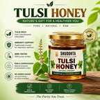 A jar of Shuddhta Tulsi Honey displayed on a wooden surface with a green and white promotional background. The jar has a gold lid and a white label with green and red text.